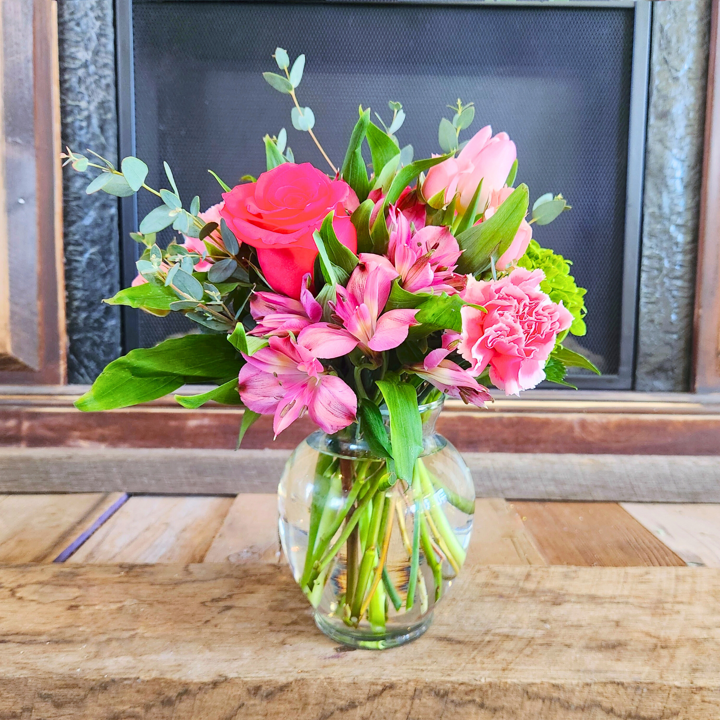 A medium-sized bouquet of pink flowers, including roses and tulips, arranged in a clear glass vase, with a fireplace in the background.