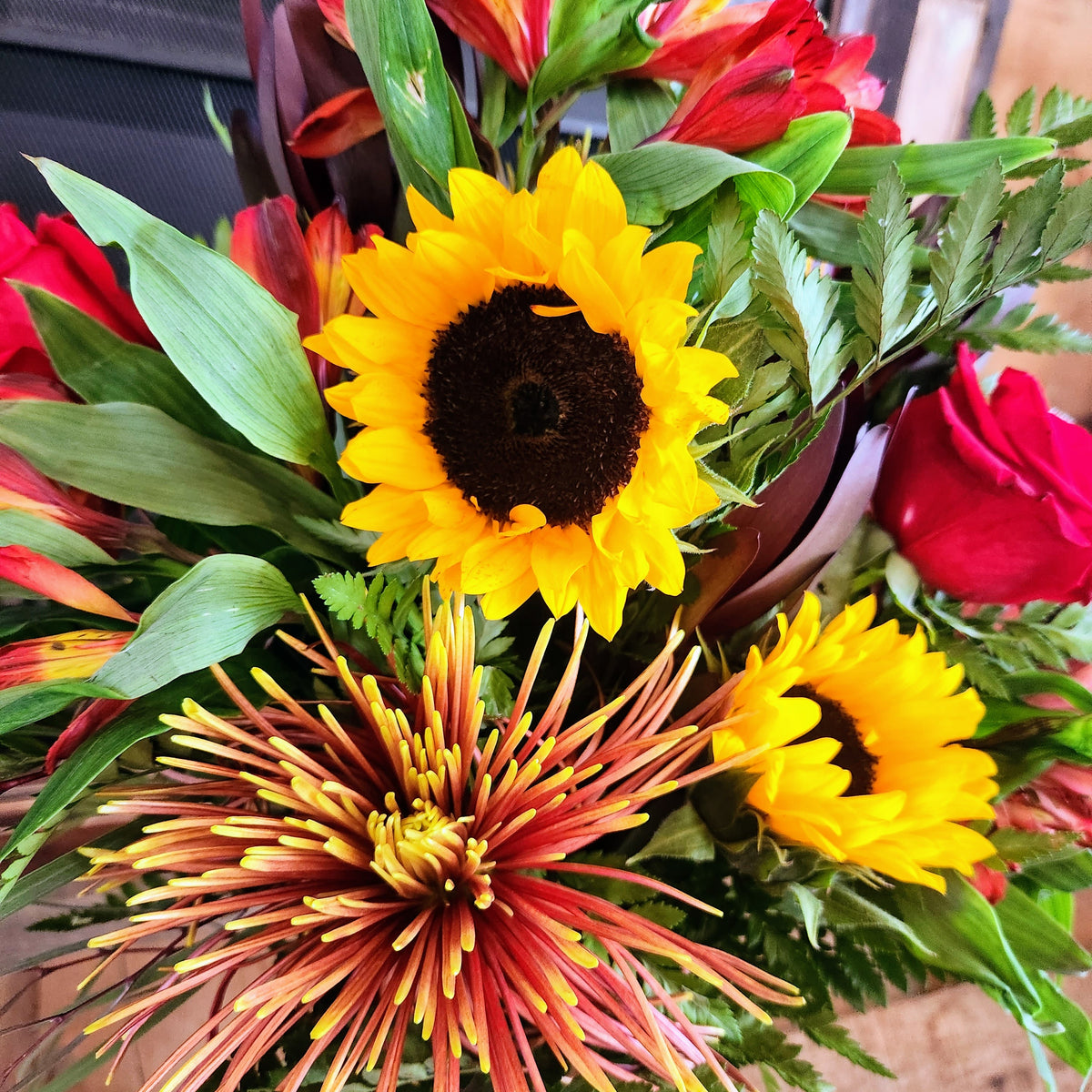 Close-up of sunflowers, red roses, fall accent flowers, and textured greenery in the Autumn Velvet Bouquet from Treasured Roots Flower Shop in Hoopeston, IL.