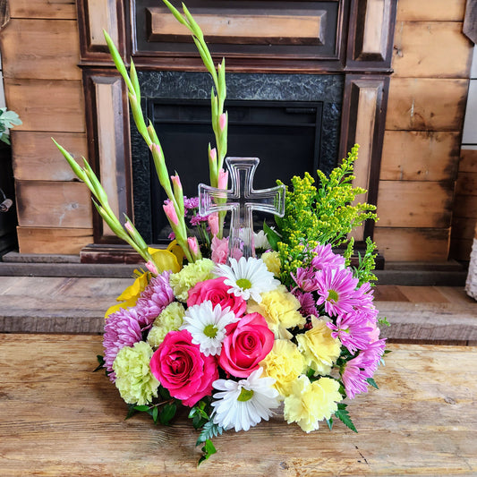 A colorful floral arrangement featuring a crystal cross, with a variety of flowers including pink roses and other plants, displayed in front of a wooden background.