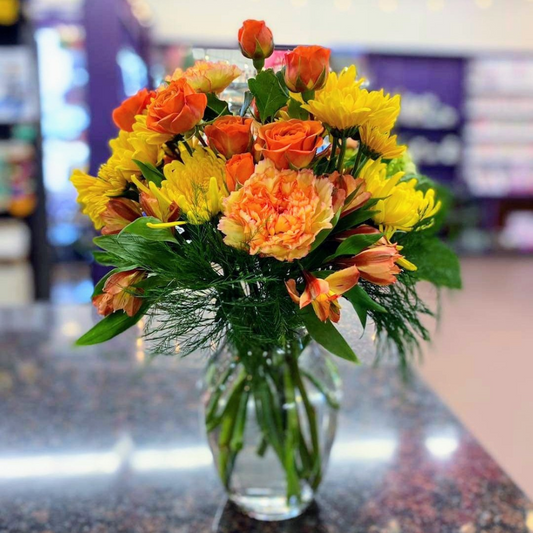 A vibrant bouquet of yellow and orange flowers including carnations, alstroemeria, daisies, and spray roses, arranged in a clear glass vase.