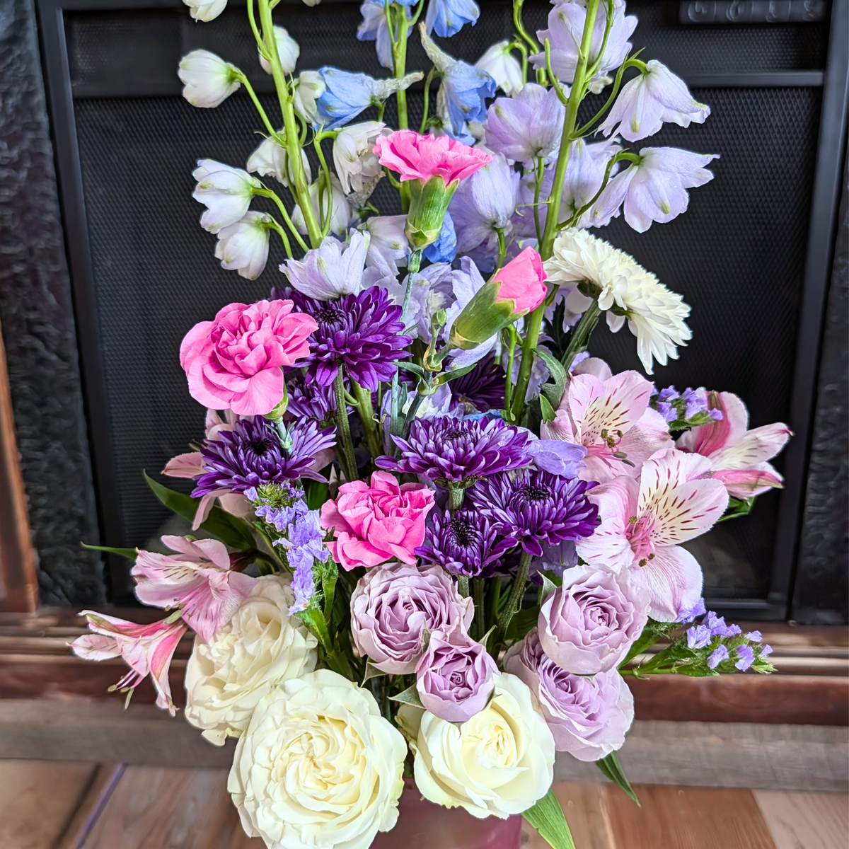 Close-up of the Cotton Candy Skies Bouquet featuring pink carnations, lavender roses, purple mums, pink alstroemeria, blue delphinium and white flowers from Treasured Roots Flower Shop in Hoopeston, IL.