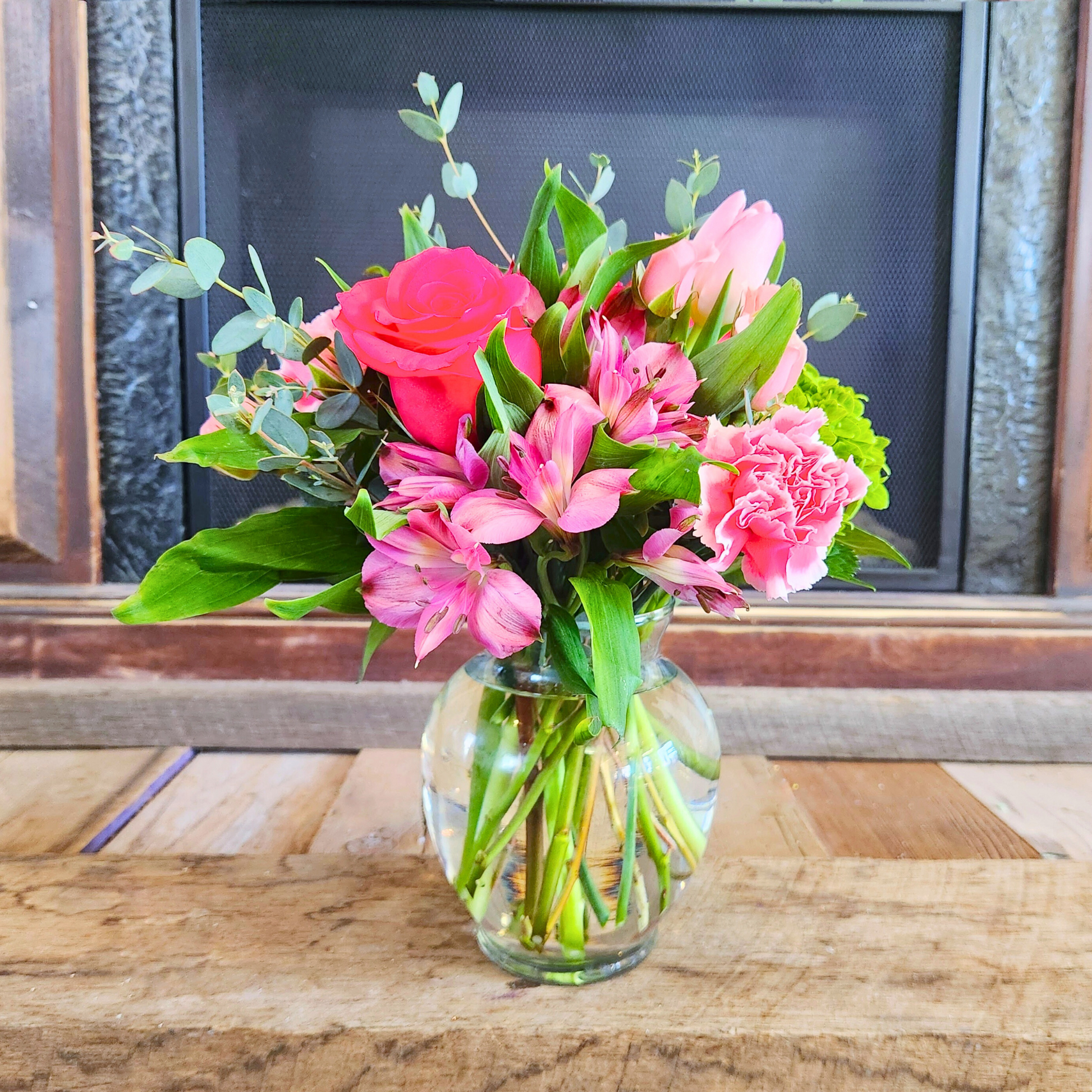 A medium-sized bouquet of pink flowers, including roses and tulips, arranged in a clear glass vase, with a fireplace in the background.