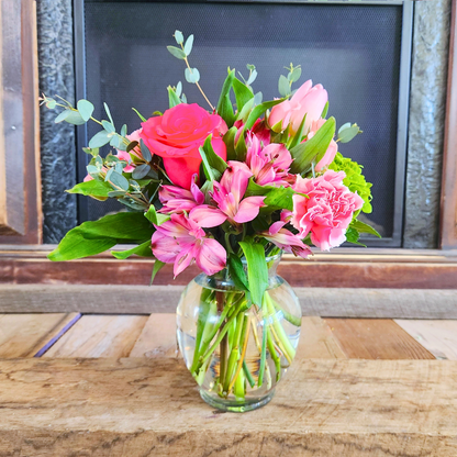 A medium-sized bouquet of pink flowers, including roses and tulips, arranged in a clear glass vase, with a fireplace in the background.