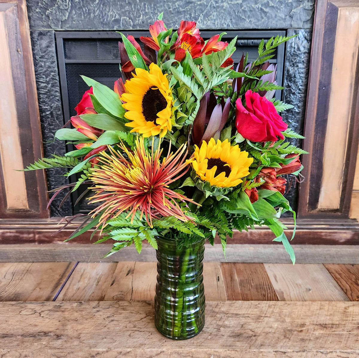 Tall Autumn Velvet fall bouquet with sunflowers, red roses, seasonal accent flowers, and lush greenery in a green glass vase from Treasured Roots Flower Shop in Hoopeston, IL.