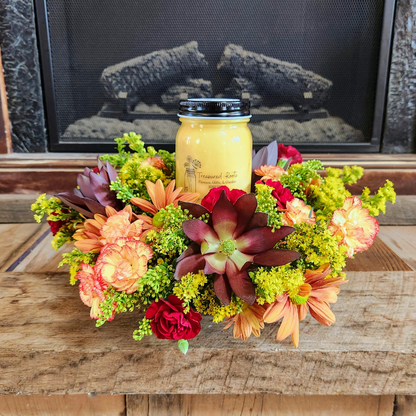 Cozy Glow fall centerpiece with orange, yellow, red, and burgundy flowers arranged around a mason jar candle from Treasured Roots Flower Shop in Hoopeston, IL.