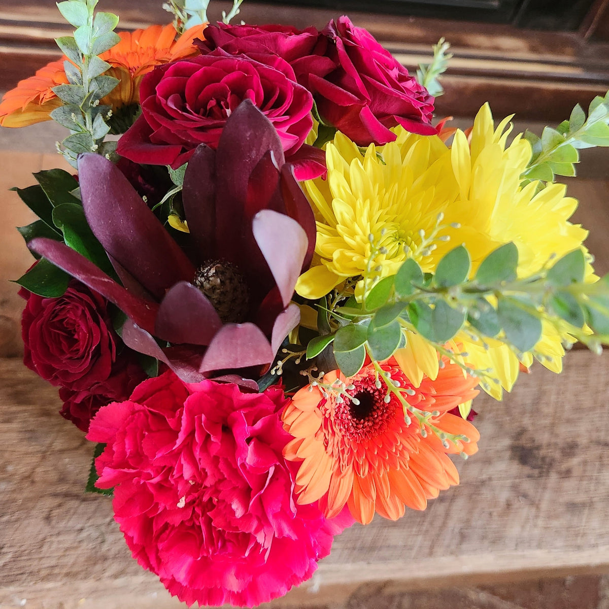 Close-up of Crimson Crush Bouquet showing red, burgundy, pink, orange, and yellow fall flowers with light eucalyptus and seasonal greenery from Treasured Roots Flower Shop in Hoopeston, IL.