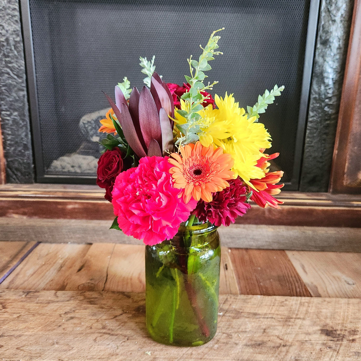 Crimson Crush fall bouquet with rich red and burgundy flowers, bright orange, yellow and pink blooms, eucalyptus, and greenery in a green fall jar vase from Treasured Roots Flower Shop in Hoopeston, IL.