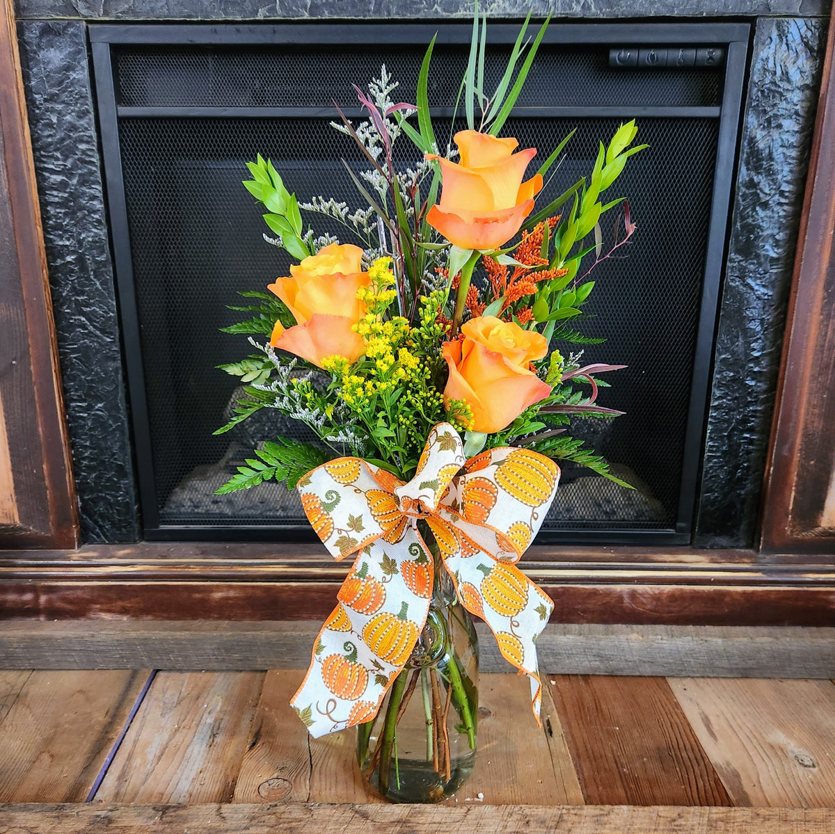 Tall Enchanted Harvest fall bouquet with three orange roses, seasonal accents, greenery, and a pumpkin-print bow in a glass vase from Treasured Roots Flower Shop in Hoopeston, IL.