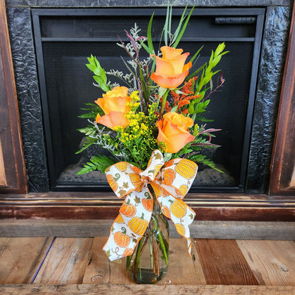 Tall Enchanted Harvest fall bouquet with three orange roses, seasonal accents, greenery, and a pumpkin-print bow in a glass vase from Treasured Roots Flower Shop in Hoopeston, IL.
