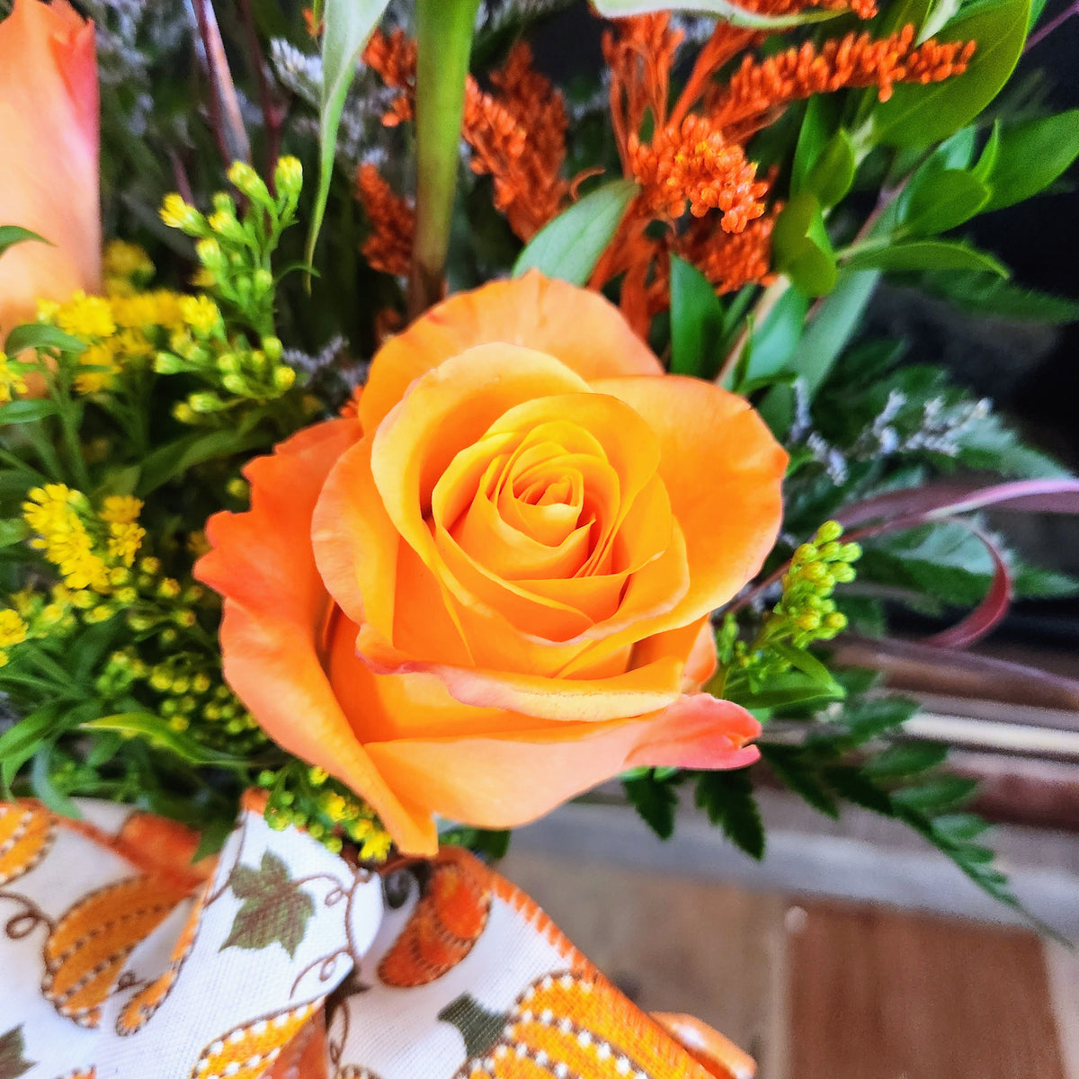 Close-up of an orange rose with coordinating fall flowers and greenery in the Enchanted Harvest Bouquet from Treasured Roots Flower Shop in Hoopeston, IL.
