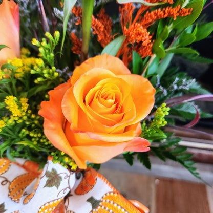 Close-up of an orange rose with coordinating fall flowers and greenery in the Enchanted Harvest Bouquet from Treasured Roots Flower Shop in Hoopeston, IL.