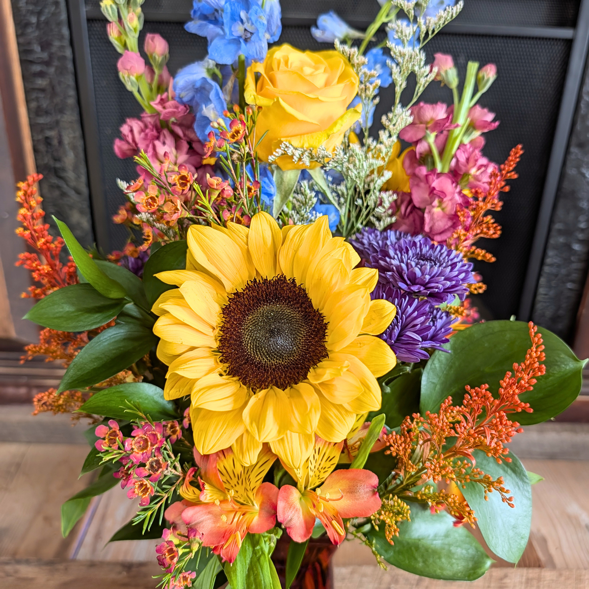 Close-up of Harvest Hues showing a sunflower, yellow rose, blue delphinium, mauve flowers, purple poms, and warm fall accents from Treasured Roots Flower Shop in Hoopeston, IL.