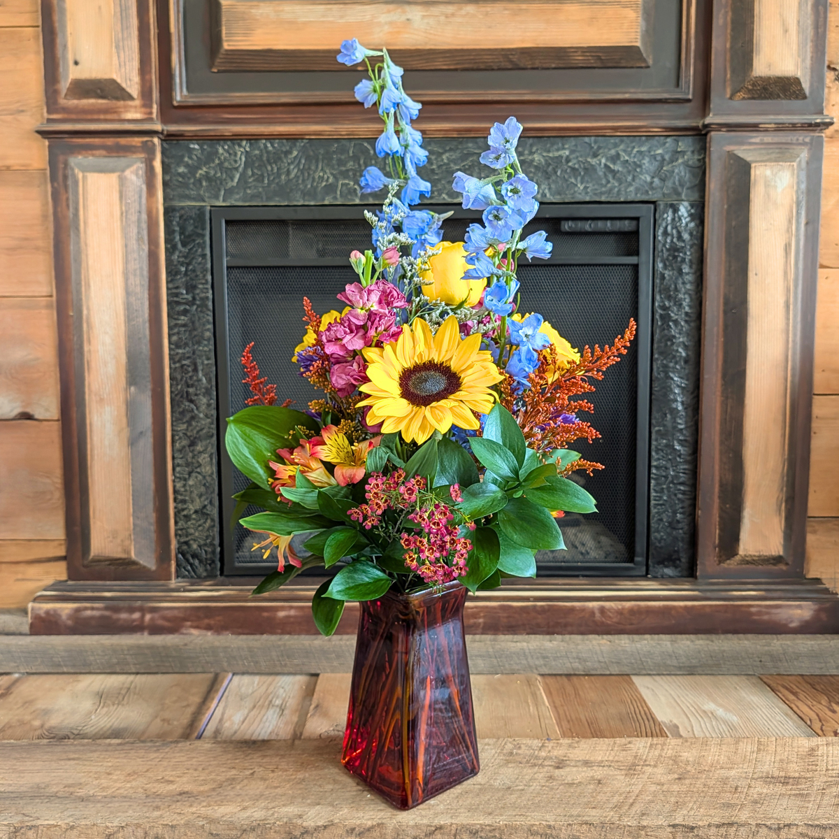 Harvest Hues tall fall bouquet with sunflowers, yellow, blue, mauve, red, and orange flowers in a red glass vase from Treasured Roots Flower Shop in Hoopeston, IL.