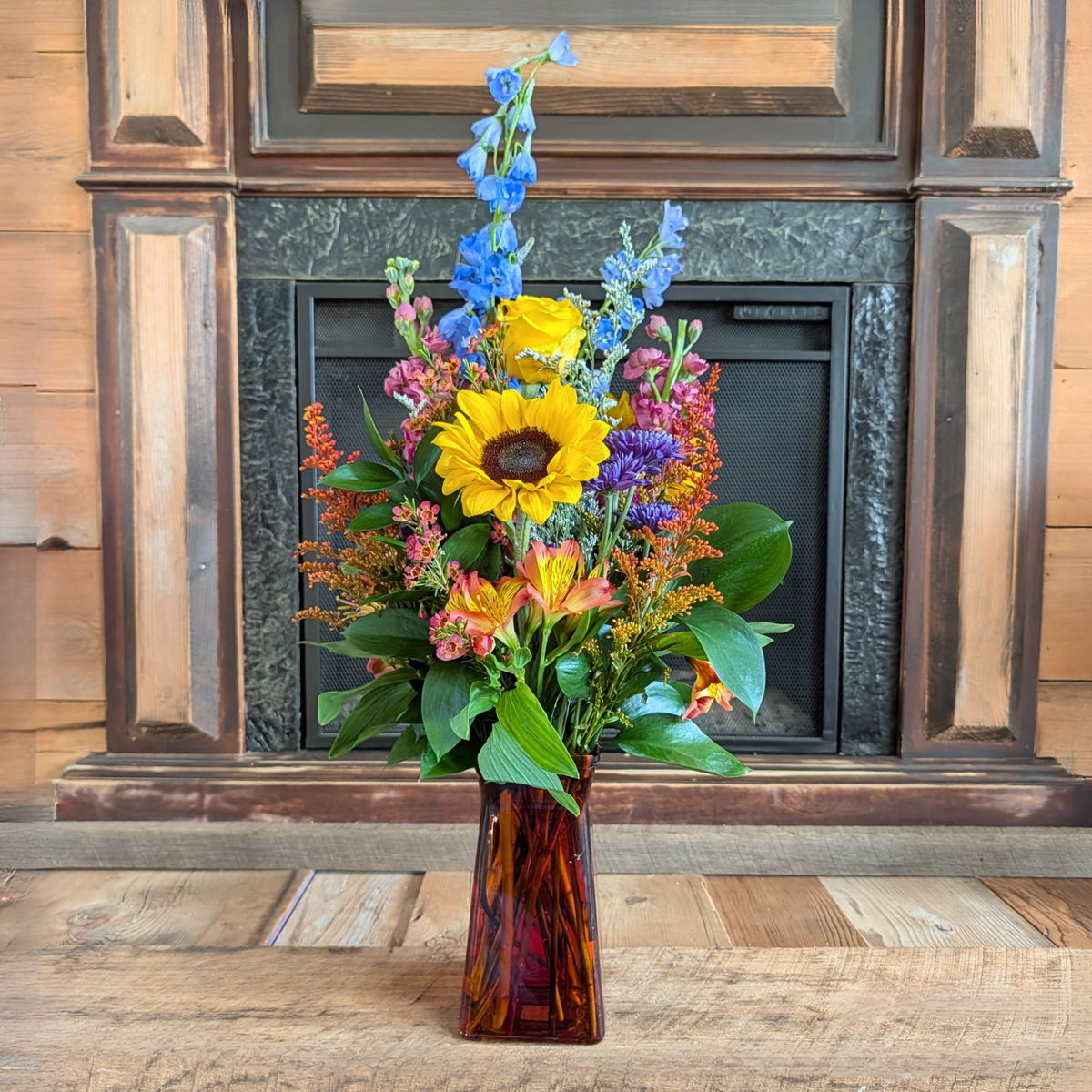 Tall Harvest Hues fall flower arrangement featuring sunflowers and colorful seasonal blooms in a red glass vase from Treasured Roots Flower Shop in Hoopeston, IL.