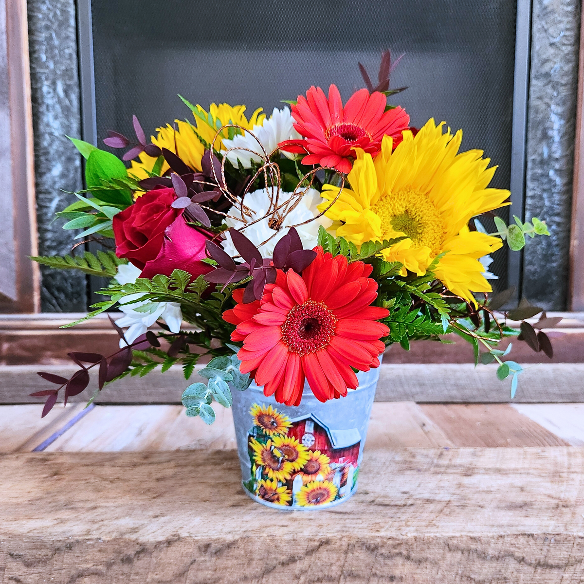 Petal to the Metal fall flower arrangement with sunflowers, red gerbera daisies, roses, white flowers, mixed greenery, and a copper wire flower in a fall-themed metal tin from Treasured Roots Flower Shop in Hoopeston, IL.