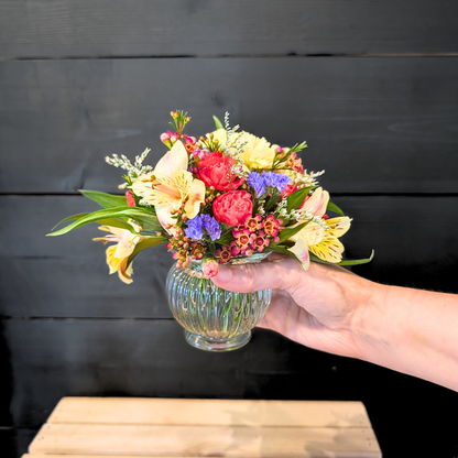 Hand holding a petite fall flower bouquet with red, yellow, purple, and cream seasonal blooms in a small clear ribbed vase.