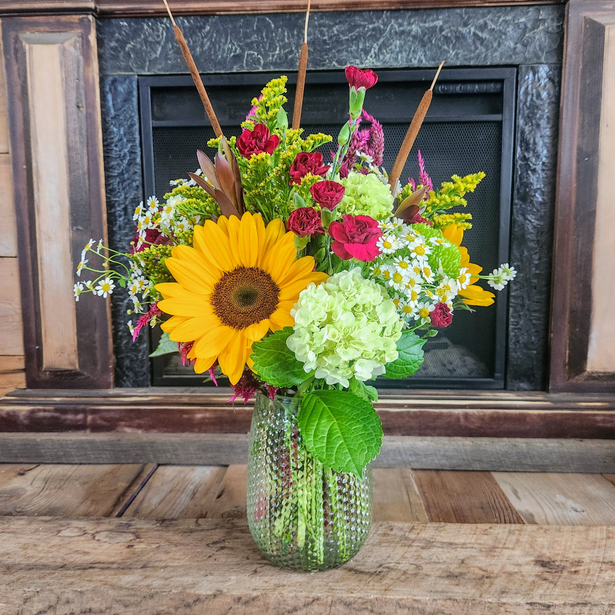 Woodland Stroll fall bouquet with yellow sunflowers, green hydrangea, burgundy flowers, purple celosia, cattails, and mixed seasonal greenery in a glass vase from Treasured Roots Flower Shop in Hoopeston, IL.
