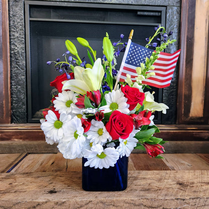 A square blue glass vase arranged with a bouquet of red, white, and blue flowers, including a small American flag, placed on a wooden surface with a fireplace in the background.