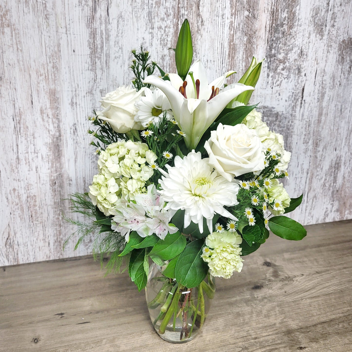 A floral arrangement featuring a white lily, white roses, and various greenery in a clear vase against a rustic wooden backdrop.