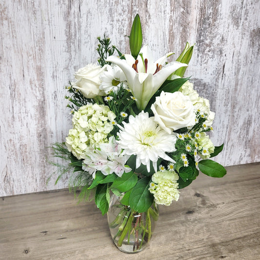 A floral arrangement featuring a white lily, white roses, and various greenery in a clear vase against a rustic wooden backdrop.