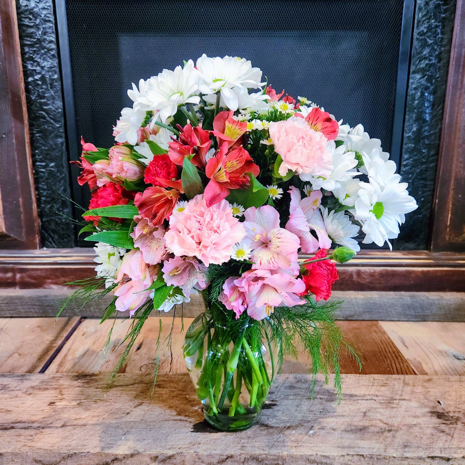 A bouquet of mixed flowers including daisies and carnations in shades of white and pink, with greenery and a vase, placed on a wooden surface in front of a fireplace.