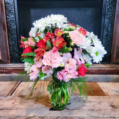 A bouquet of mixed flowers including daisies and carnations in shades of white and pink, with greenery and a vase, placed on a wooden surface in front of a fireplace.